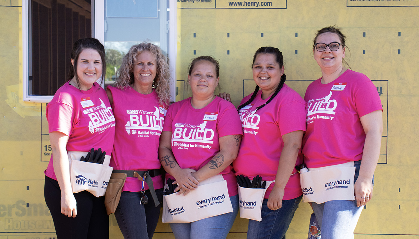 Five women standing together, smiling in pink shirts and tool belts in front of a new home construction.